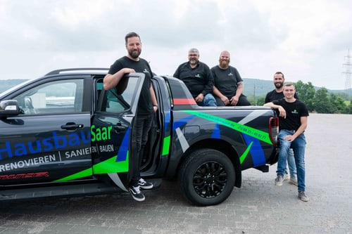 Fünf Männer posieren bei einem schwarzen Pickup-Truck mit "Hausbau Saar" und "Renovieren Sanieren Bauen" an der Seite.